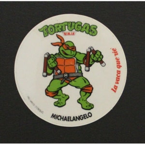 Op de afbeelding: Een ronde sticker met een cartoon Teenage Mutant Ninja Turtle, Michelangelo, die nunchaku's vasthoudt. De sticker is wit met rode, oranje, groene en zwarte accenten. De tekst "Tortugas Ninja" en "La vaca que ríe" staat op de sticker gedrukt.