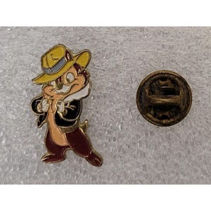 May include: A gold-coloured pin featuring a cartoon chipmunk wearing a yellow hat and a brown jacket. The chipmunk is standing with its arms crossed and has a white fur collar. The pin has a gold-coloured back with a safety pin closure.