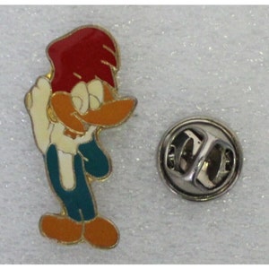 Op de afbeelding: Een cartoon pin van Woody Woodpecker met een rode hoed, een oranje snavel, een wit lichaam, een tealblauwe overall en oranje schoenen. De pin heeft een zilveren metalen achterkant met een veiligheidsspeld.