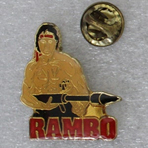 May include: A gold-colored pin featuring a cartoon image of John Rambo from the movie "Rambo". He is holding a large gun and wearing a red headband. The word "RAMBO" is written in red letters below the image.