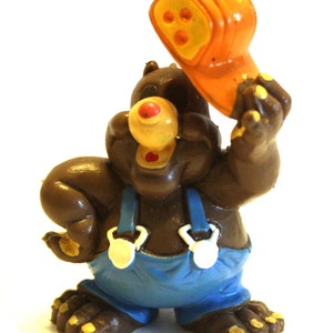 May include: A brown cartoon bear figurine wearing blue overalls with white clasps, holding an orange object with three holes. The bear has a yellow nose and red mouth, with yellow accents on its feet and hands.