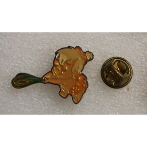 Puede incluir: Un pin de metal dorado con un personaje de dibujos animados sosteniendo un garrote verde. El personaje lleva un taparrabos marrón y tiene la piel naranja. El pin tiene una parte posterior dorada con un pasador de seguridad.