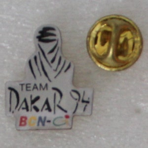 May include: A white enamel pin with a black and white illustration of a person wearing a headscarf. The pin says "TEAM DAKAR 94 BCN-CI" in black text.