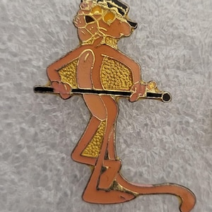 Può includere: Spilla vintage in smalto raffigurante la Pantera Rosa in posa mentre cammina, con un bastone nero. La spilla ha uno sfondo strutturato color oro ed è bordata in oro. La Pantera Rosa è raffigurata in tonalità rosa e arancione, con un cappello nero.