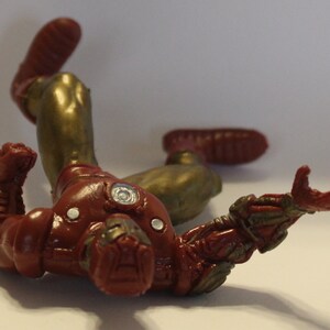 May include: A red and gold action figure of Iron Man lying on its back with its arms outstretched. The figure is made of plastic and has a detailed paint job.