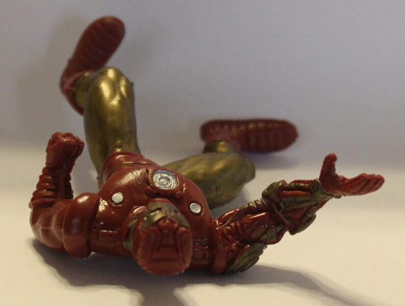 May include: A red and gold action figure of Iron Man lying on its back with its arms outstretched. The figure is made of plastic and has a detailed paint job.