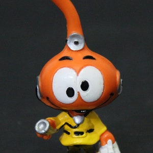 Puede incluir: Figurita de plástico naranja y amarilla de un personaje de dibujos animados con una nariz larga naranja, ojos grandes blancos y una chaqueta amarilla. El personaje sostiene una pequeña bolsa médica blanca y una linterna.