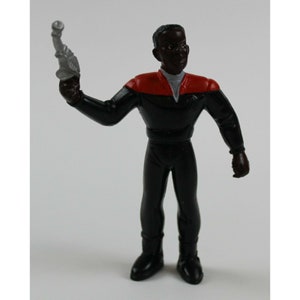 Op de afbeelding: Een zwart action figure van een Star Trek-personage dat een zwart uniform draagt met rode accenten. De figuur houdt een zilveren phaser in zijn rechterhand.