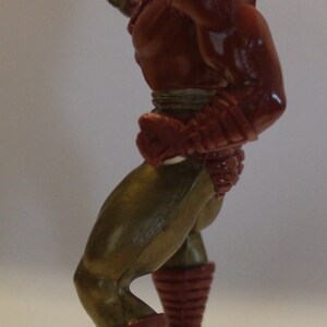 May include: A red and gold action figure of a superhero with a raised arm and a closed fist. The figure is posed in a dynamic stance with one leg forward and the other leg back. The figure has a detailed sculpt and paint job.
