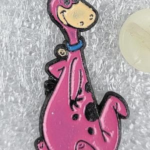 Puede incluir: Un pin de dinosaurio de dibujos animados rosa con contornos negros y manchas negras. El dinosaurio tiene una cara amarilla, un collar azul y un mechón de pelo negro. El pin está sobre un fondo plateado texturizado.