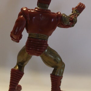 May include: A red and gold plastic action figure of a robot with a raised arm. The figure has a detailed design with a segmented body and a helmet.