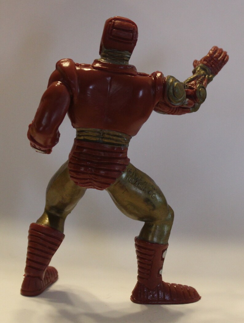 May include: A red and gold plastic action figure of a robot with a raised arm. The figure has a detailed design with a segmented body and a helmet.