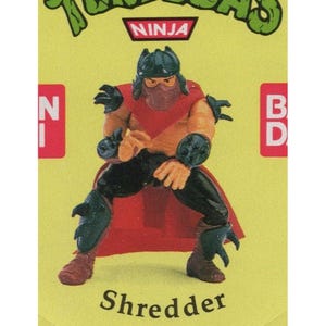 May include: A Shredder action figure from the Teenage Mutant Ninja Turtles franchise. The figure is posed on a yellow background and features a red cape, black armor, and a purple mask. The word "Shredder" is printed at the bottom.