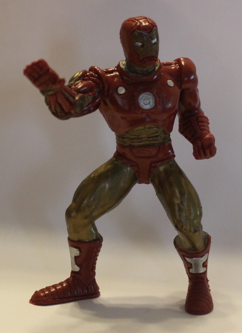 May include: A red and gold plastic action figure of Iron Man, a superhero from Marvel Comics. The figure is posed in a fighting stance with one arm raised and the other fist clenched. The figure has a red and gold suit with a silver chest plate and a red helmet with a gold faceplate.