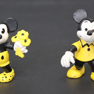 May include: Two vintage plastic Mickey Mouse figurines. One Mickey is wearing yellow shorts and holding a bouquet of yellow flowers. The other Mickey is wearing a yellow polo shirt.
