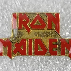 Puede incluir: Un pin metálico dorado con las palabras "IRON MAIDEN" en letras rojas. El pin tiene forma rectangular con un aspecto ligeramente desgastado, y es probablemente un artículo de colección.