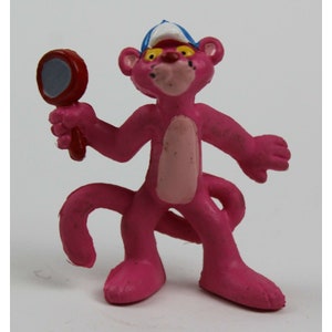 May include: Pink Panther figurine wearing a blue baseball cap and holding a red magnifying glass. The figurine is made of plastic and has a pink and white color scheme.