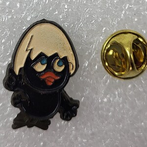May include: A black and tan enamel pin featuring a cartoon chick hatching from an egg. The chick has blue eyes and a red beak. A gold-colored pin back is next to the chick.