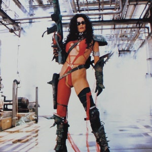 Op de afbeelding: Een vrouw in een rood en zwart kostuum houdt een groot geweer vast. Ze draagt een rood en zwart bodypak met een zwarte riem en een zwarte helm. Ze staat in een fabriekomgeving met pijpen en andere industriële apparatuur op de achtergrond.