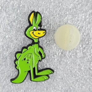 Puede incluir: Un pin de dinosaurio de dibujos animados verde con detalles amarillos y un contorno negro. Junto a él, un pequeño objeto redondo de color blanquecino. Un accesorio divertido y caprichoso.