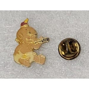May include: A gold-toned pin featuring a cartoon baby with a crown, holding a flute. The baby is wearing a diaper. A separate gold-toned pin back is also visible.