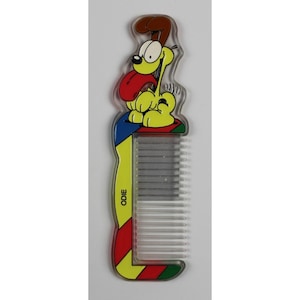 May include: A clear plastic comb featuring the cartoon character Odie. Odie is yellow with a red tongue and brown ears. The comb's handle is yellow with the name "ODIE" printed on it, and has a colorful striped design.