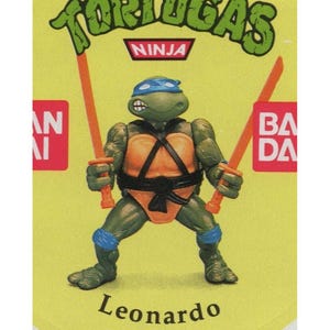 Op de afbeelding: Een groene Teenage Mutant Ninja Turtle-actiefiguur met een blauwe bandana, een oranje schild en een zwarte riem. De figuur houdt twee oranje katanas vast. De figuur staat op een gele achtergrond met de woorden "Tortugas Ninja" en "Leonardo" eronder.