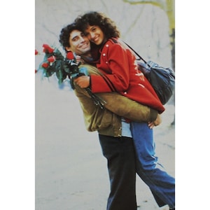 May include: A man in a brown jacket holds a woman in a red jacket with black stripes. The woman is wearing blue jeans and the man is wearing blue jeans and black pants. The man is holding a bouquet of red roses.