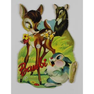 May include: A vintage children's book cover featuring Bambi the deer, a rabbit, and a doe. The cover is illustrated in a cartoon style with bright colors and a green background. The title "Bambi" is written in red letters.