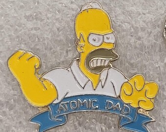 May include: A yellow enamel pin featuring Homer Simpson with an angry expression. He is wearing a white shirt and the pin has a blue banner that reads "ATOMIC DAD".