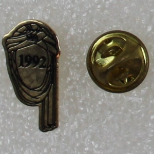 May include: Gold-colored pin with a design of a stylized head with the year 1992 in the center. The pin has a back with a safety clasp.