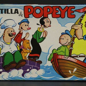 May include: Vintage Popeye the Sailor cartoon box featuring various characters, including Popeye, Olive Oyl, and Bluto. The box has a blue background with the words "IMPRENTILLA de POPEYE" in red.