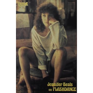 May include: A vintage movie poster featuring Jennifer Beals from the film Flashdance. The poster has a warm, sepia tone. Jennifer Beals is seated, wearing a light gray sweater. The text "Jennifer Beals en Flashdance" is at the bottom.
