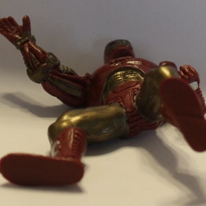 May include: A red and gold action figure of a superhero lying on its back with one arm outstretched. The figure has a metallic finish and is made of plastic.