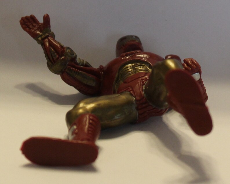 May include: A red and gold action figure of a superhero lying on its back with one arm outstretched. The figure has a metallic finish and is made of plastic.