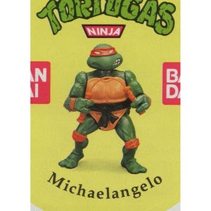 Op de afbeelding: Een groene Teenage Mutant Ninja Turtles actionfiguur met een oranje schild, een rode bandana en een zwarte riem. De figuur houdt een nunchaku in zijn rechterhand. De figuur staat op een gele achtergrond met de woorden "Teenage Mutant Ninja Turtles" en "Ninja" erboven gedrukt. De naam van de figuur, "Michelangelo", is eronder gedrukt.