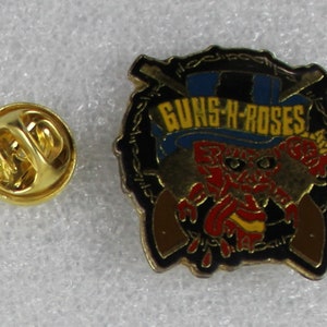 May include: A gold-colored pin with a black background featuring a cartoon character wearing a top hat with the text "GUNS N' ROSES".