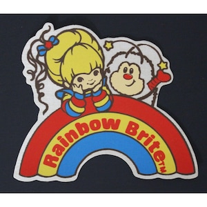 May include: A yellow-haired girl with a red bow sits on a rainbow with the words "Rainbow Brite" in yellow and red letters. A white furry creature with a red bow sits next to her.