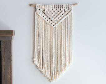 May include: A white macrame wall hanging with a wooden dowel rod, a light wood nightstand with a drawer, and a small potted succulent plant in a white geometric planter.