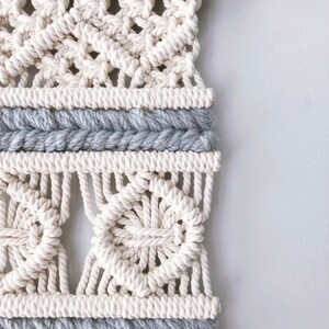May include: A white macrame wall hanging with a gray and white pattern. The hanging is made with thick rope and has a natural wood dowel at the top.