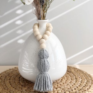Peut inclure: Un pompon gris suspendu à un vase en céramique blanche avec une guirlande de perles. Le vase est posé sur un dessous de plat en paille tressée.