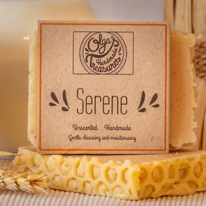 Puede incluir: Una barra de jabón artesanal sin perfume etiquetada como "Serene" con el texto "Olga's Treasures" en la etiqueta. El jabón está sobre una superficie con patrón de panal de abeja, con avena y tallos de trigo en el fondo.