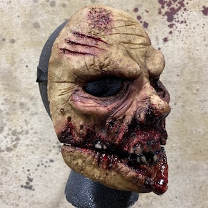 Peut inclure: Un masque de zombie détaillé avec une apparence réaliste et macabre. Le masque est fait d'un matériau beige et présente une grande bouche ouverte avec des dents visibles, des éclaboussures de sang et une blessure réaliste sur le front.