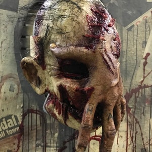 Puede incluir: Una máscara de terror realista con una mano cubriendo la cara. La máscara está hecha de látex y tiene un aspecto sangriento y grotesco. La mano está detallada con dedos y uñas.