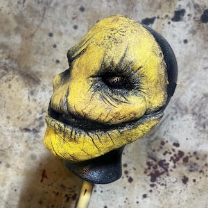 May include: A yellow and black Halloween mask with a sinister, carved face. The mask has a textured, aged appearance, with deep lines and a menacing grin. The mask is mounted on a black base with a curved handle.