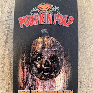 May include: A black and orange enamel pin featuring a scary pumpkin with a carved face and green eyes. The pin is on a black card with the text "Pumpkin Pulp" and "www.pumpkinpulp.com".