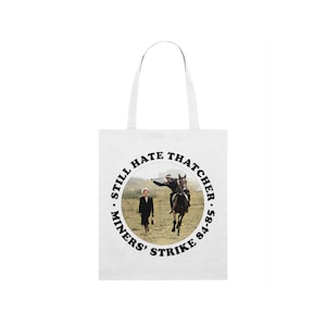 Puede incluir: Bolsa de tela blanca con un gráfico circular negro. El gráfico presenta una foto de Margaret Thatcher montando a caballo, con una mujer caminando detrás de ella. El texto alrededor de la foto dice "Still Hate Thatcher - Miners' Strike 84-85."