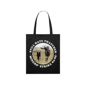Puede incluir: Bolso tote negro con un diseño gráfico circular que dice "Still Hate Thatcher Miners' Strike 84-85". El diseño muestra a una persona a caballo y a otra caminando en un campo. El bolso tiene dos asas negras.