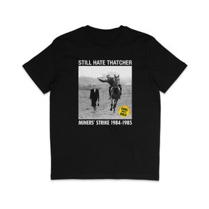 Puede incluir: Camiseta negra con un gráfico blanco de Margaret Thatcher a caballo, una persona caminando detrás de ella y un cartel amarillo que dice "Coal Not Dole". El texto "Still Hate Thatcher" y "Miners' Strike 1984-1985" también está impreso en la camiseta.