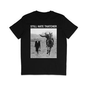 Puede incluir: Camiseta negra y blanca con el texto "STILL HATE THATCHER" impreso en blanco. La camiseta presenta una foto en blanco y negro de Margaret Thatcher caminando lejos de una persona que monta a caballo.
